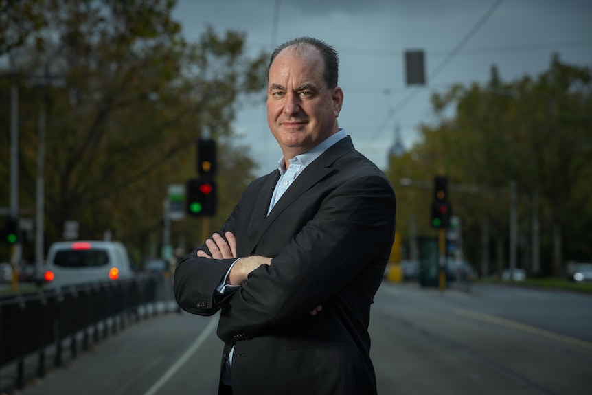 a man in a business suit stand in the middle of a road, with a grey melbourne sky in the background.