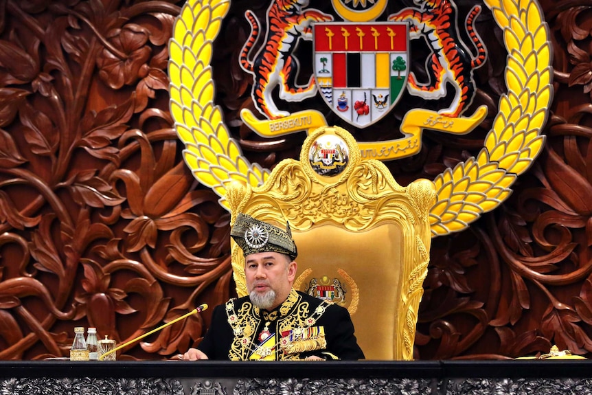 A man sitting on a gold throne in a traditional outfit in front of a large emblem.