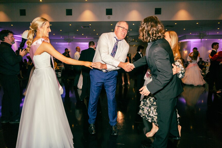 A man and a teenager shake hands on a dance floor next to a girl in a formal white dress.