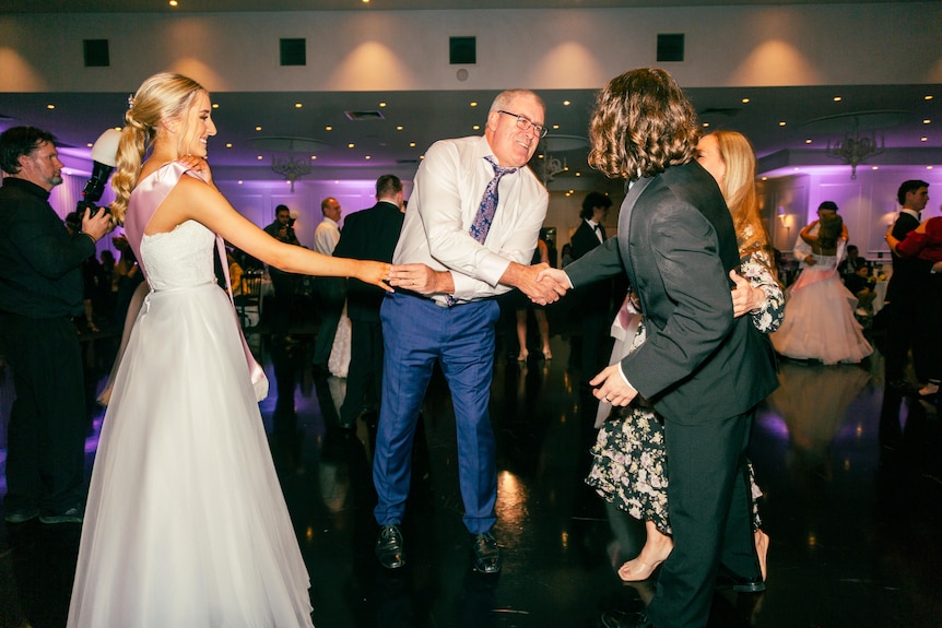 A man and a teenager shake hands on a dance floor next to a girl in a formal white dress.