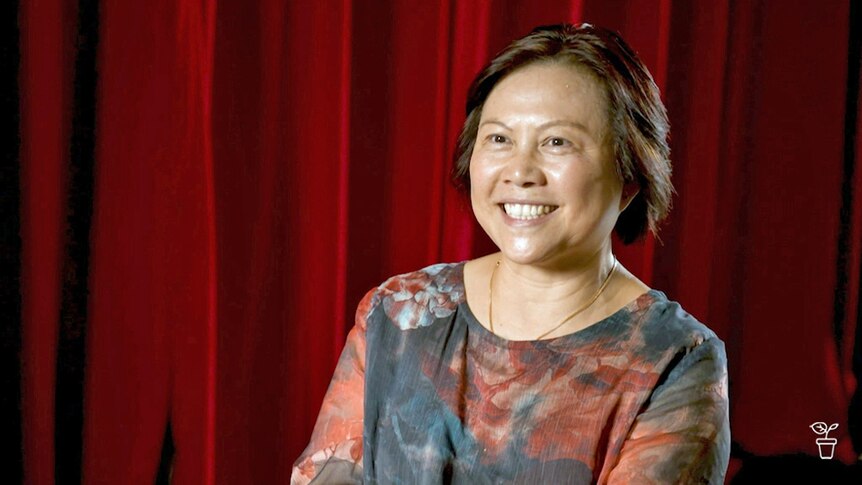 Woman smiling, sitting in front of a dark red curtain.