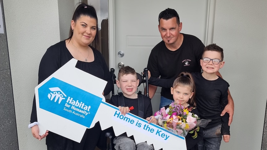 Family photo of Mum and Dad with boy sitting on walker, little girl with flowers and boy with glasses  in front of house door