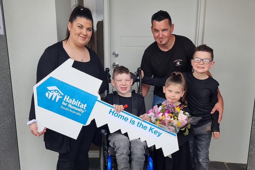 Family photo of Mum and Dad with boy sitting on walker, little girl with flowers and boy with glasses  in front of house door