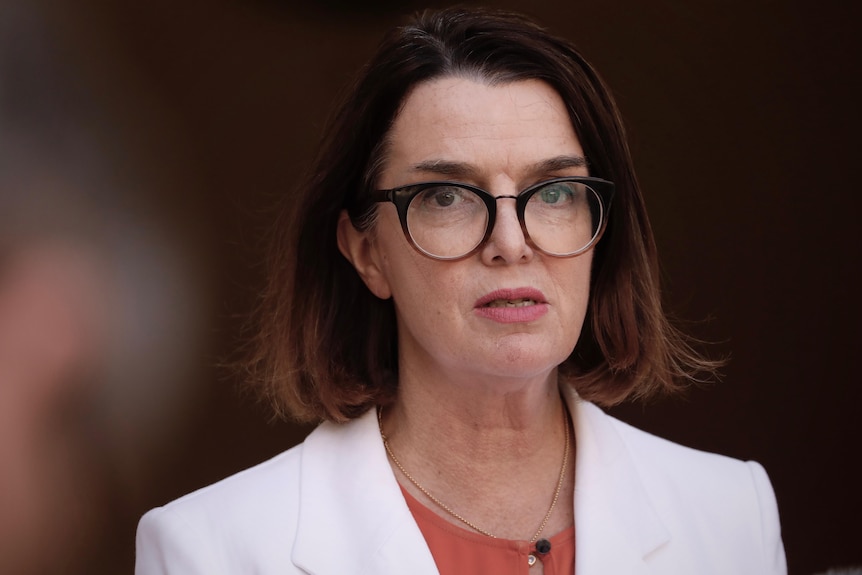 Un headshot d'une femme avec des cheveux bruns aux épaules et des lunettes portant un blazer blanc