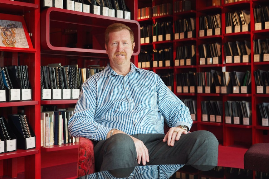 A man sits in a chair surrounded by red bookshelves and bound books.