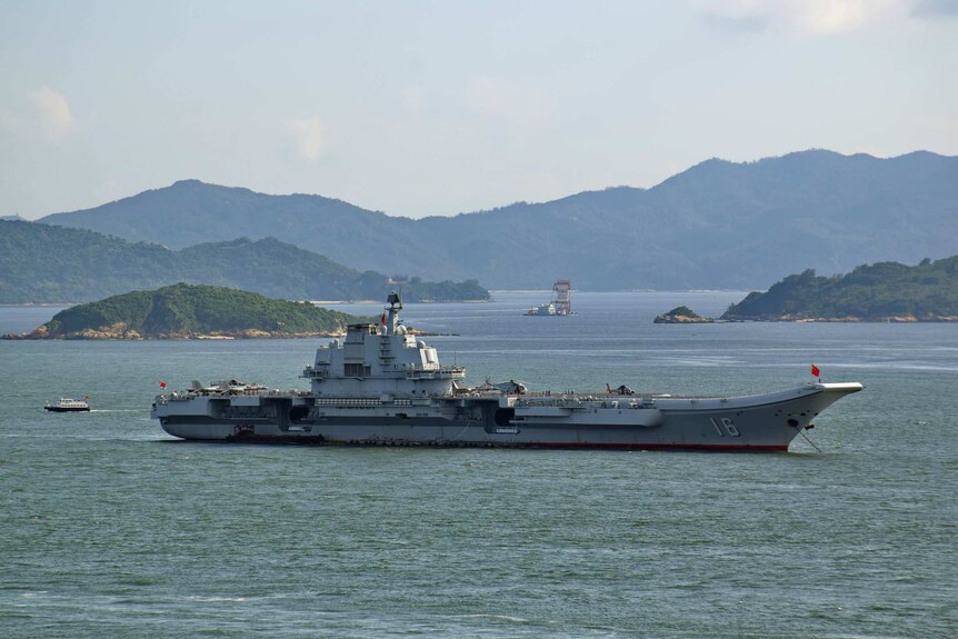 A large aircraft carrier sits in the middle of a harbour with various inlets and mountainous peninsulas shown in the distance.