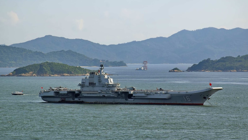 A large aircraft carrier sits in the middle of a harbour with various inlets and mountainous peninsulas shown in the distance.