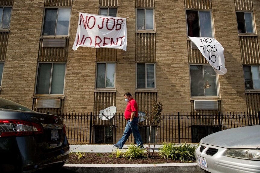 signs hang from a windows of an apartment block reading no job no rent as a man with a mask walks past