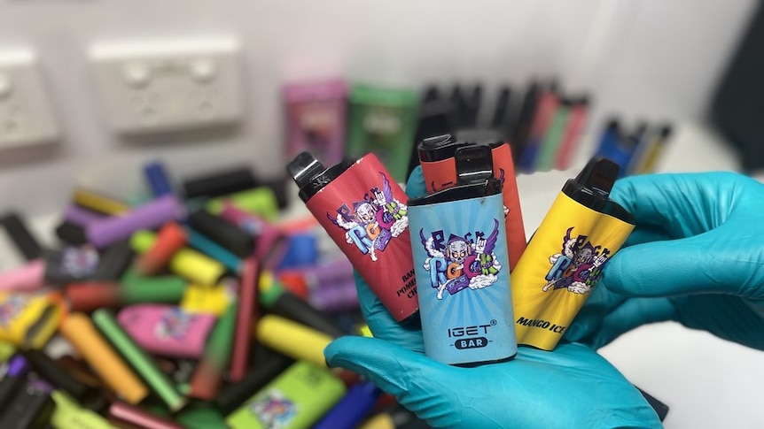 Gloved hands hold a selection of vapes above a bin of multi-coloured objects.