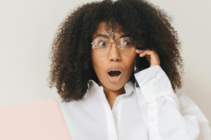 A shocked-looking woman with dark curly hair holds a laptop in one hand, a phone to her ear in the other