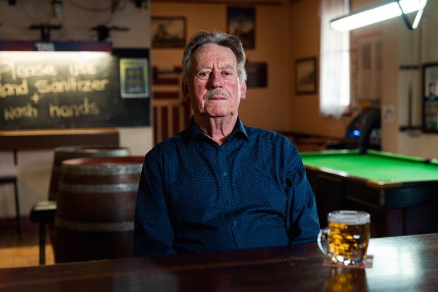 Claye wearing a dark blue shirt sitting at the front bar of a pub, with a glass of beer on the counter and a pool table in back.