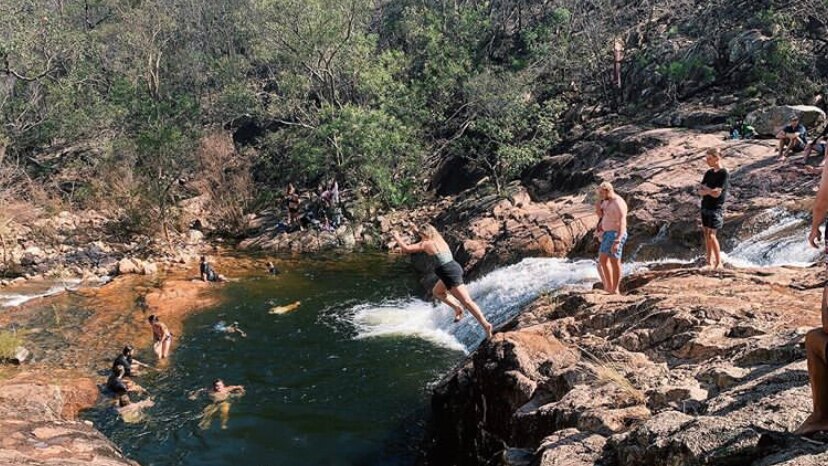 About a dozen people gathered at a water hole in the Australian bush. A young woman leaps off a rock into the water.