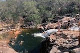 About a dozen people gathered at a water hole in the Australian bush. A young woman leaps off a rock into the water.