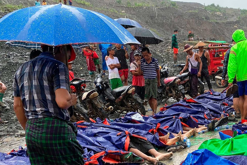 People gather near a row of bodies covered in tarp on the side of a muddy mountain.