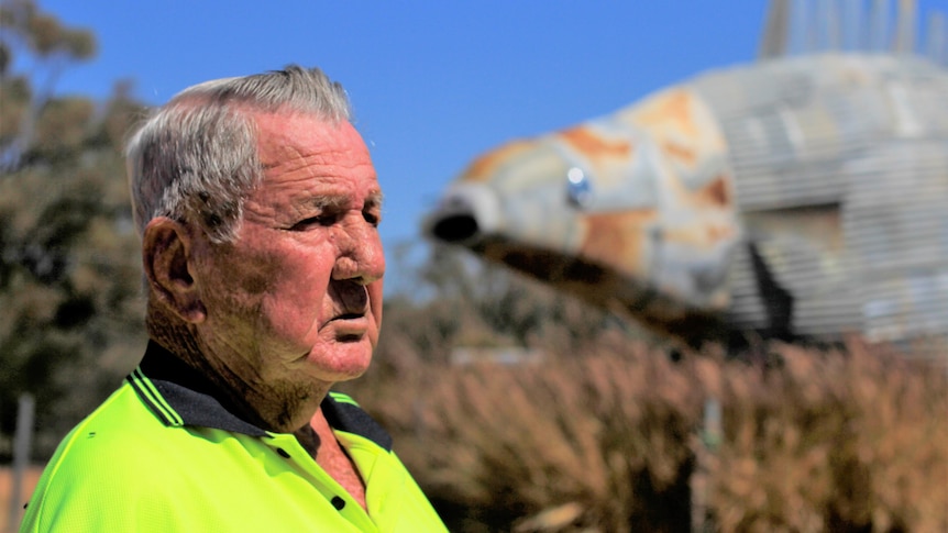 An older man in a yellow hi-vis shirt stands in front of a giant metal fish.