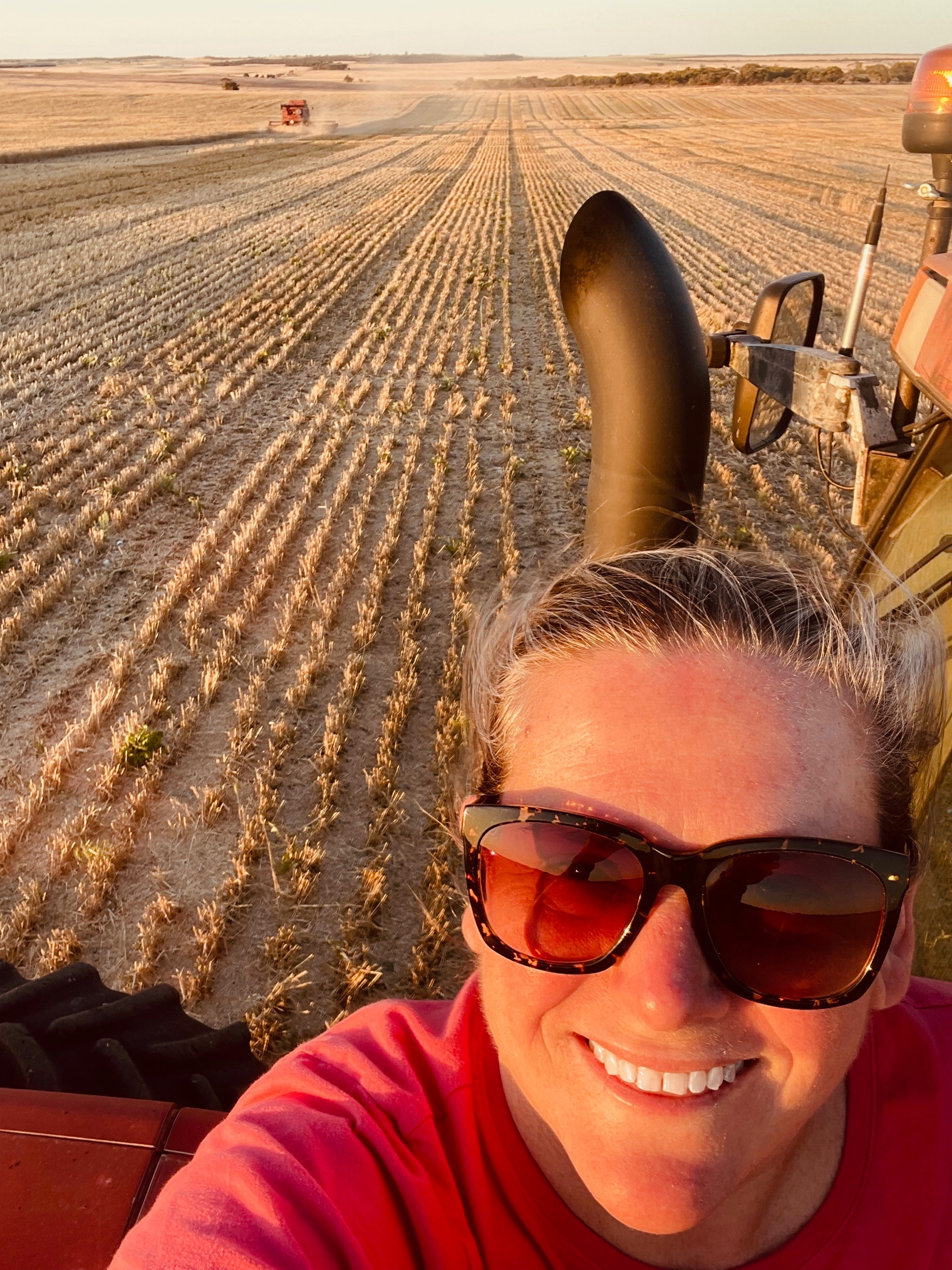 Una mujer sonriente sentada en un tractor con un prado seco y una cosecha de trigo cortada al fondo.