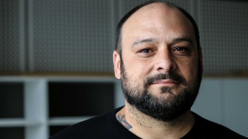 Close-up shot of man in black shirt, with red tattoo visible at his neck, short black hair and neutral expression.