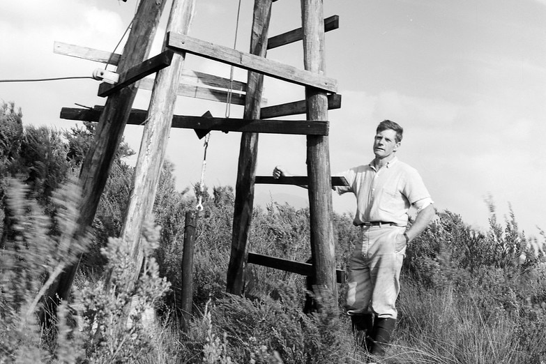 Black and white photo of man outdoors next to a wooden structure 