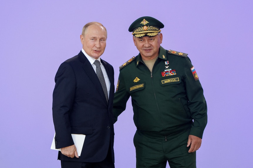 Vladimir Putin and Sergei Shoigu put their arms around each other in front of a pink wall
