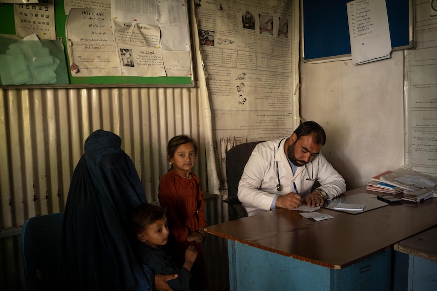 A Middle Eastern doctor in white coat writes on notepad as a covered woman and her daugther look on in office