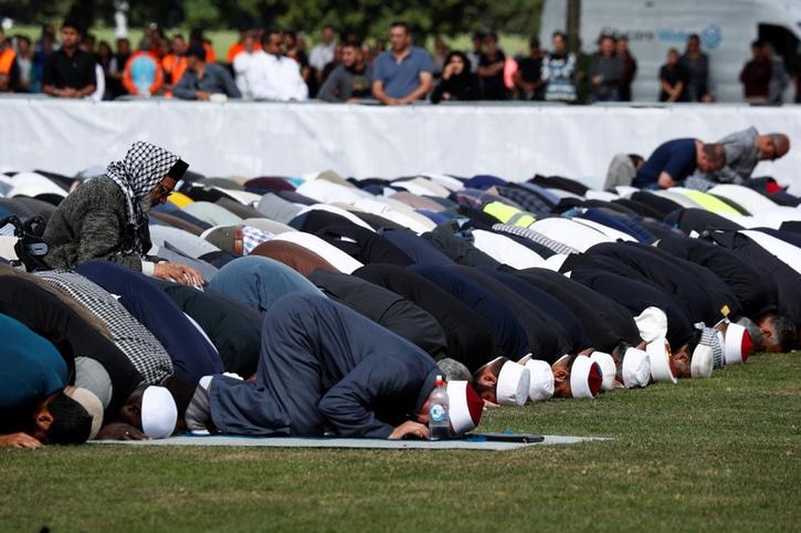 Four rows of people are seen kneeling as they pray with their heads to the ground on mats sitting on grass.