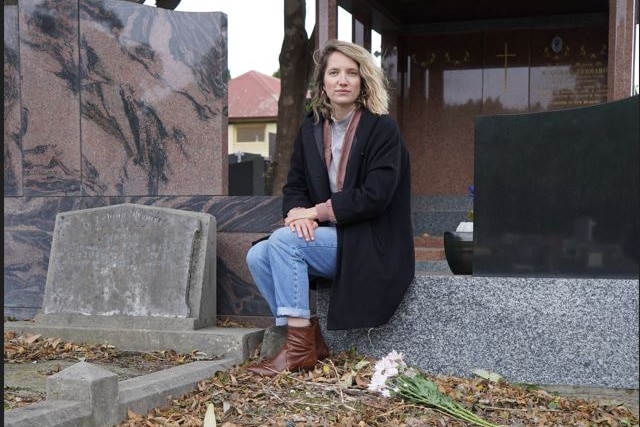 A woman wearing a black coat and blue jeans sits at the headstone of a bare grave