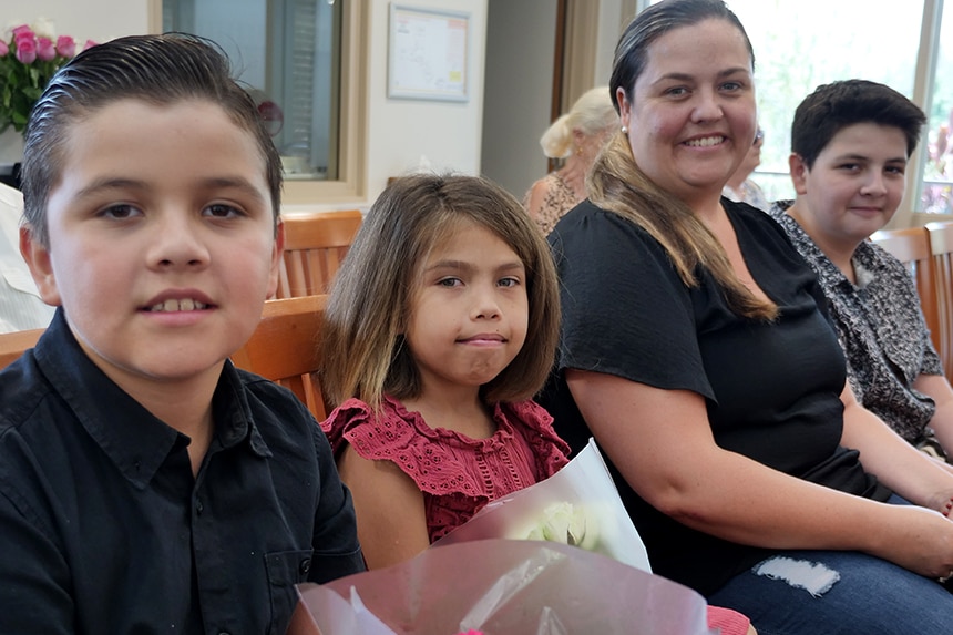A woman in a black shirt is seated with her children. They are all smiling.