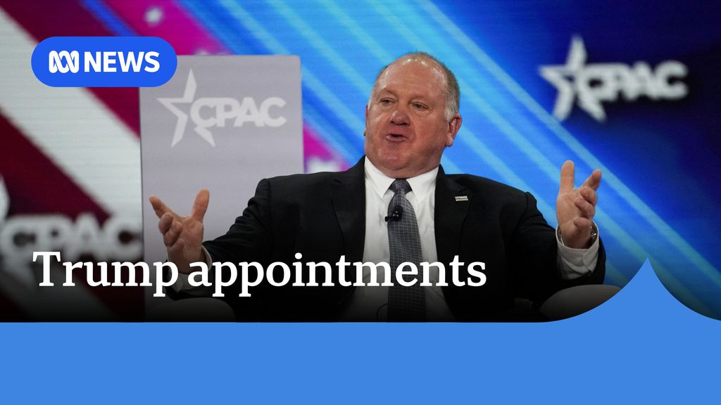 Trump appointments: Man sits in a chair with arms open gesticulating in front of bright banner.