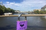A purple padlock with 'Efti loves Jodie' written on it hangs from a wire, city and river in background.