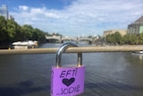 A purple padlock with 'Efti loves Jodie' written on it hangs from a wire, city and river in background.