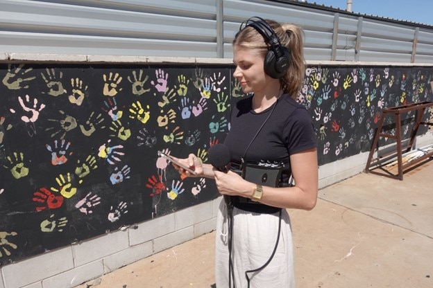 Woman holding microphone and audio equipment, wearing headphones, standing in front of wall with handprints.