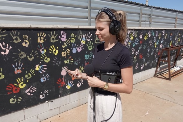 Woman holding microphone and audio equipment, wearing headphones, standing in front of wall with handprints.