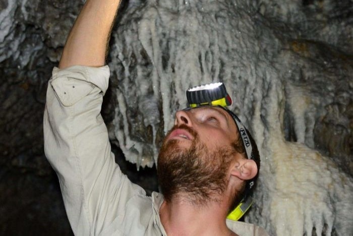 Surrounded by the wet, smooth, white walls of a cave, a man wearing headlamp looks up reaching to touch rock above his head.