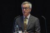 man in suit and glasses at a desk behind a microphone