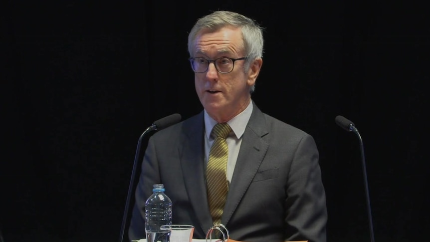 man in suit and glasses at a desk behind a microphone