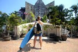 A woman with a surfboard stands in front of a memorial with flags and plants.