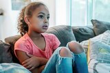 Girl sits on couch with shocked, worried lock on her face as she watches TV