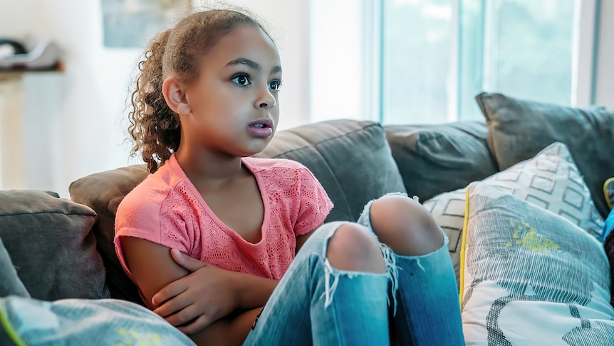 Girl sits on couch with shocked, worried lock on her face as she watches TV