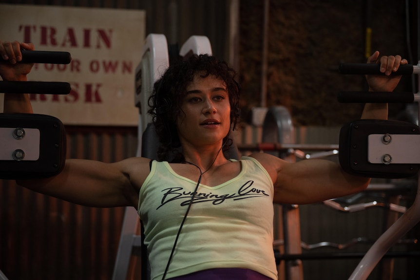 A film still of Katy O'Brian using an arm workout machine at the gym. She is wearing a singlet and headphones.