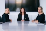 Three women sit at the end of a long table in a boardroom