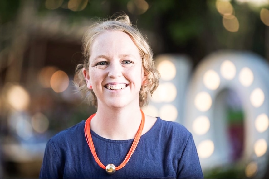 A close up of a smiling woman with blonde hair wearing a blue top and an orange necklace