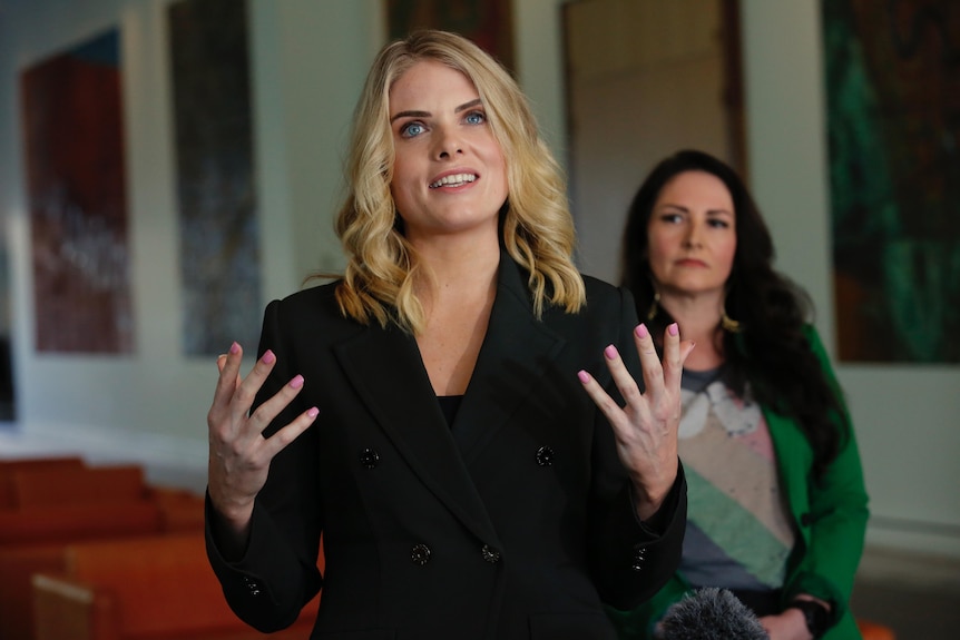 A woman with shoulder length blonde hair wearing a black blazer mid-sentence gesturing with her hands