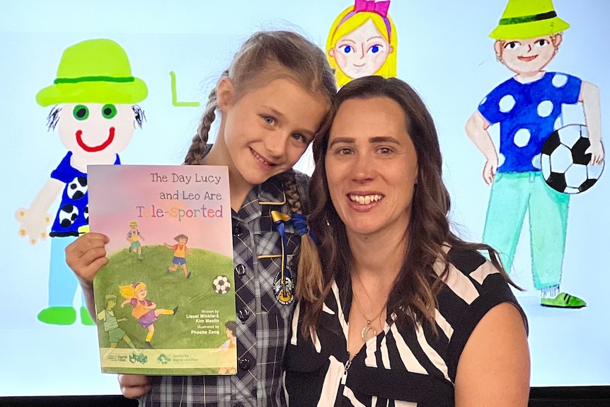 A smiling girl in a primary school uniform holds up a book while her dark-haired mum kneels beside her.