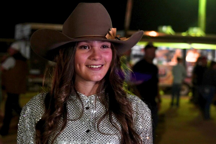 a young cowgirl with long brown hair wearing a cowboy hat and a sparkly silver button-up shirt