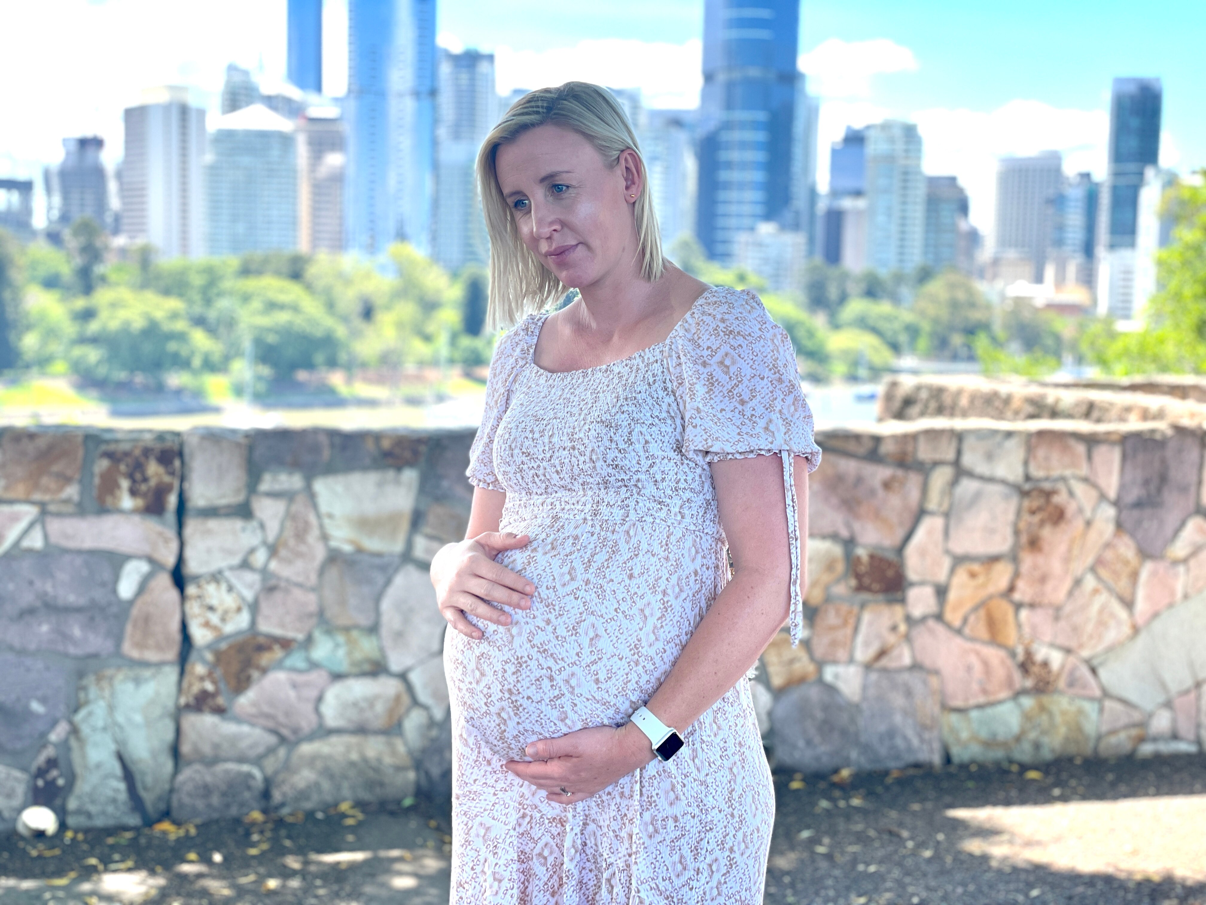 A woman in a dress holds her baby bump with Brisbane city in the background.