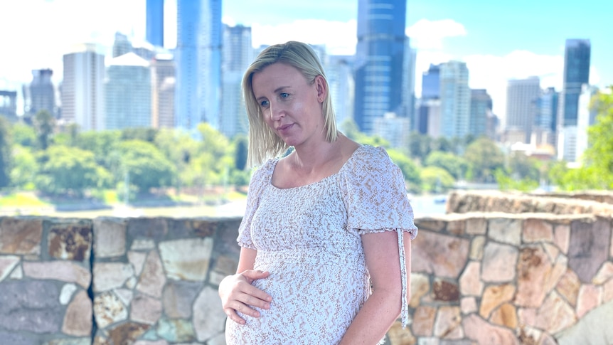 A woman in a dress holds her baby bump with Brisbane city in the background.
