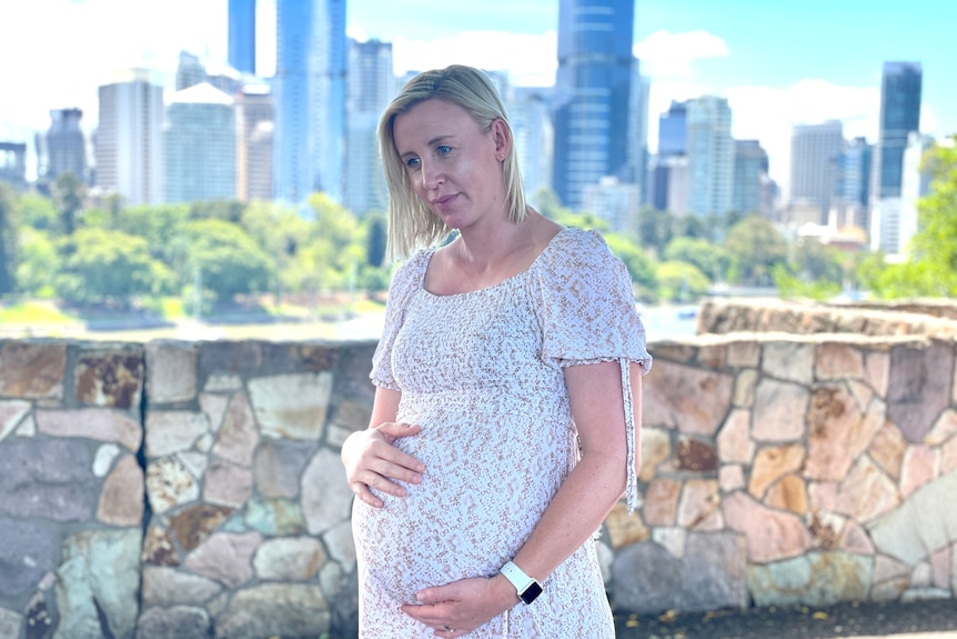 A woman in a dress holds her baby bump with Brisbane city in the background.