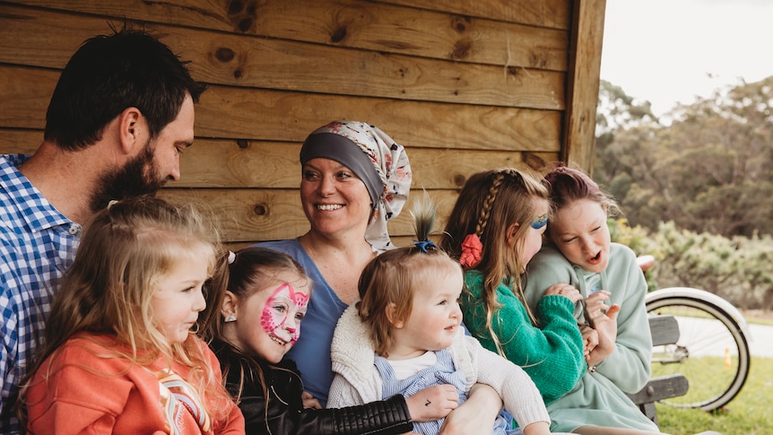 Claire and her husband are sitting down on a bench with their five young daughters on their lap in front of a wooden barn