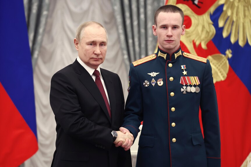 A suited politician shakes hands with a uniformed soldier while they stand before Russian flags.