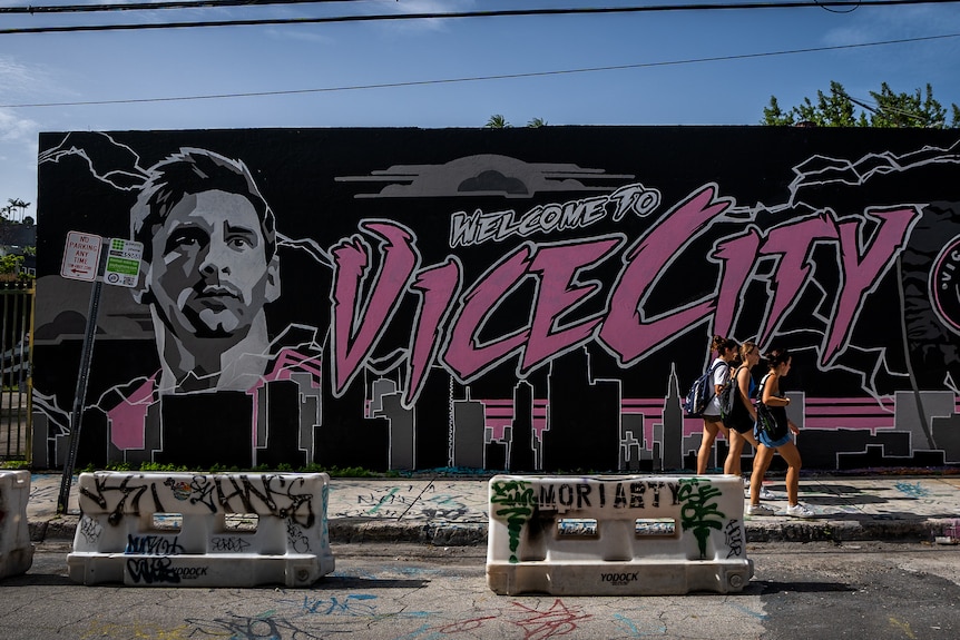 Two women walk past a mural that says WELCOME TO VICE CITY with a portrait of Lionel Messi